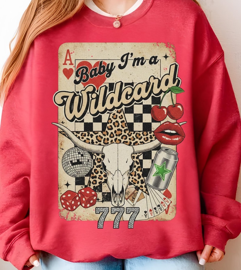 May include: Red sweatshirt with a graphic design featuring a bull skull, cherries, dice, a disco ball, and the text "Baby I'm a Wildcard". The design includes a leopard print star and a can with a green star.