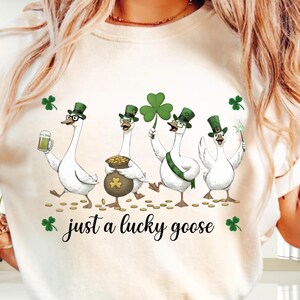 Puede incluir: Camiseta color crema con cuatro gansos de dibujos animados celebrando el D&iacute;a de San Patricio. Los gansos llevan sombreros de copa verdes y sostienen una cerveza, una olla de oro, un tr&eacute;bol y una varita m&aacute;gica. El texto "just a lucky goose" est&aacute; debajo.