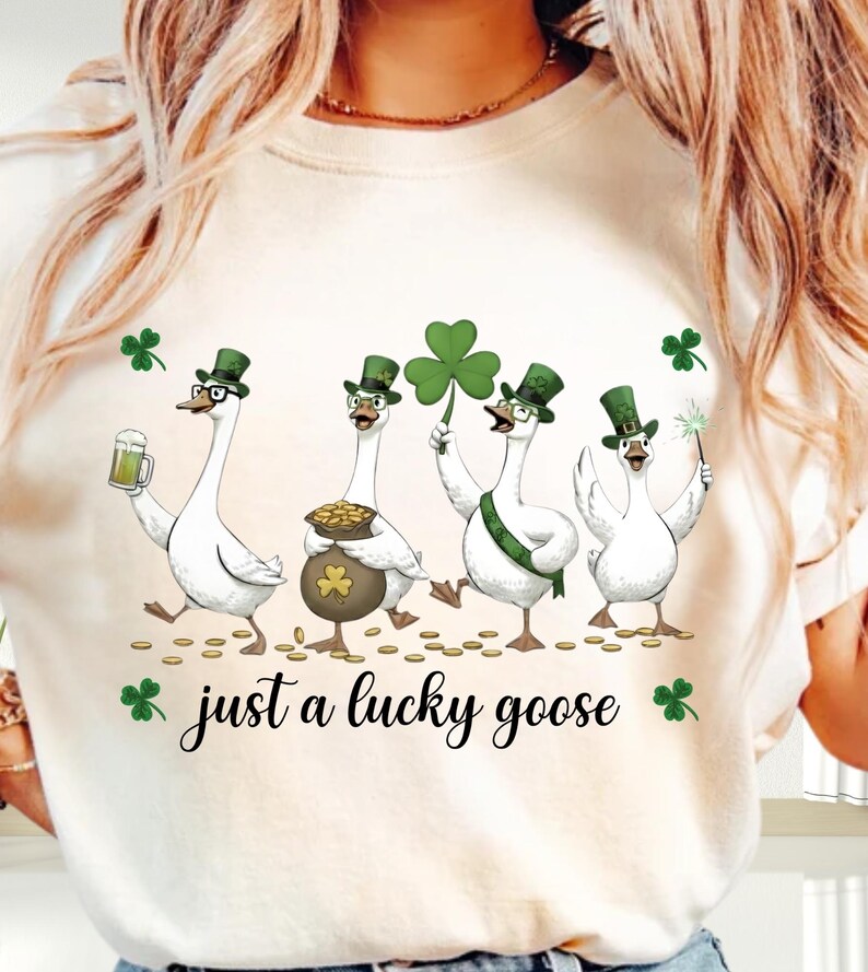 Puede incluir: Camiseta color crema con cuatro gansos de dibujos animados celebrando el D&iacute;a de San Patricio. Los gansos llevan sombreros de copa verdes y sostienen una cerveza, una olla de oro, un tr&eacute;bol y una varita m&aacute;gica. El texto "just a lucky goose" est&aacute; debajo.