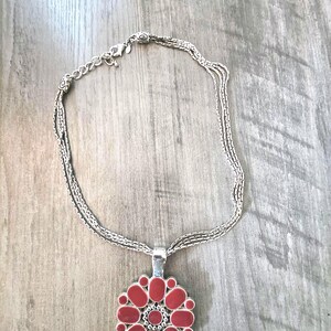 May include: A multi-strand silver-tone necklace featuring a circular pendant with a flower-like design. The pendant is composed of small, oval-shaped, reddish-orange enamel inlays arranged in a radial pattern around a central beaded silver circle.