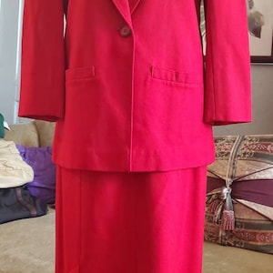 Può includere: Un tailleur rosso composto da giacca e gonna. La giacca ha una chiusura con un solo bottone e due tasche. La gonna è lunga fino al ginocchio.