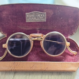 May include: Vintage round-lens spectacles with translucent frames resting inside a brown case lined with dark purple velvet. The case features a metal plaque reading W. H. WILLIAMS, M.D. EYE, EAR, NOSE AND THROAT MIDDLETOWN, OHIO.