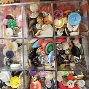 Op de afbeelding: Een doorzichtige plastic organizer gevuld met een verscheidenheid aan kleurrijke knopen. De knopen zijn in verschillende vormen, maten en kleuren, waaronder rood, blauw, geel en wit. Sommige knopen hebben ingewikkelde ontwerpen.
