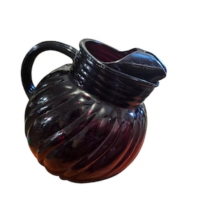 Puede incluir: Una jarra de vidrio de color morado oscuro con un cuerpo redondeado y un diseño de espiral. La jarra tiene un asa y un pico.