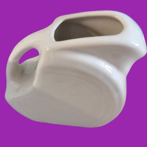 Può includere: Un creamer o un brocca in ceramica bianca con una forma arrotondata e leggermente curva. Il creamer ha un manico e una parte superiore larga e aperta.