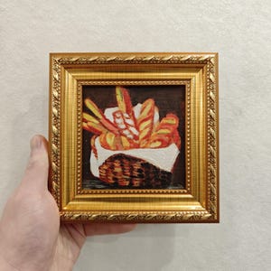 May include: A small, square oil painting in a gold frame depicts a basket of bread. The painting features various types of bread in shades of yellow, orange, and red, set against a dark brown background. The frame has a detailed, ornate design.
