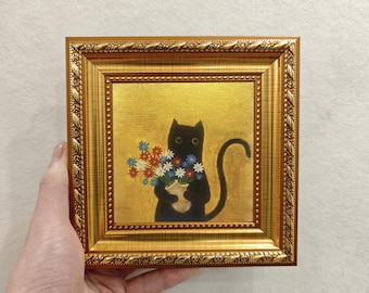 May include: A framed painting of a black cat holding a vase of colorful flowers. The artwork is set against a gold background and encased in a gold ornate frame. The painting is held by a hand.