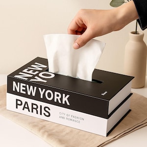 Puede incluir: Caja de pañuelos negra y blanca con las palabras "NEW YORK" y "PARIS" impresas. La caja es rectangular y tiene una ranura en la parte superior para dispensar pañuelos. El texto "CITY OF FASHION AND ROMANCE" también está impreso en la caja.