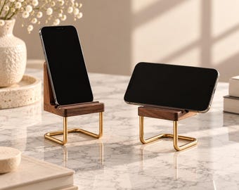 May include: Two mobile phone stands featuring dark brown walnut wood cradles supported by polished gold-tone metal frames. One stand holds a smartphone in a vertical orientation, while the other holds a smartphone in a horizontal orientation.