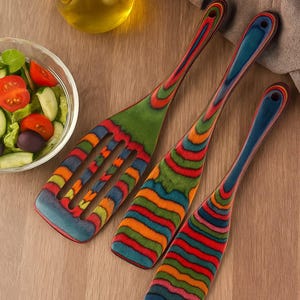 May include: A set of three colorful wooden kitchen utensils. The set includes a slotted spatula and two solid spatulas, all with vibrant rainbow-colored handles and heads. The utensils are displayed on a wooden surface, suggesting a kitchen or dining setting.