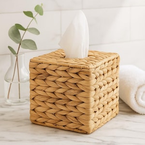 Puede incluir: Un dispensador de pañuelos cuadrado hecho de fibras naturales tejidas. La caja es de color marrón claro con una superficie texturizada y una pila de pañuelos blancos que emergen de la parte superior. Una toalla blanca enrollada y un jarrón con vegetación están en el fondo.