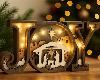 May include: Wooden Christmas decoration spelling "JOY", with a nativity scene inside the "O". The letters are illuminated with warm white lights. A Christmas tree with bokeh lights is in the background. The decoration is on a wooden surface.