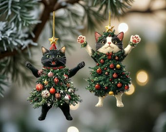 May include: Two cat-shaped Christmas ornaments. One is black, the other is black and white. Both are dressed as Christmas trees, adorned with red and gold ornaments, and wear star-topped hats. They hang from gold strings against a blurred green background.