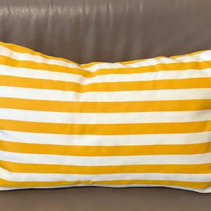 Könnte beinhalten: Rechteckiges Kissen mit horizontalen gelb-weißen Streifen. Das Kissen besteht aus Stoff und liegt auf einer braunen Oberfläche. Es ist ein dekorativer Artikel für ein Bett oder Sofa.