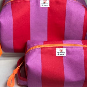 Könnte beinhalten: Zwei gestreifte Kosmetiktaschen in Rot und Lila mit orangefarbenen Reißverschlüssen und Zuglaschen. Die Taschen tragen ein Etikett mit der Aufschrift "by Maison de Maron". Ein lebendiges und farbenfrohes Accessoire.