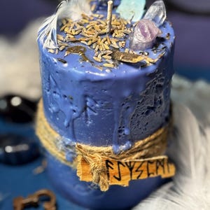Könnte beinhalten: Eine blaue Kerze mit rustikaler Ästhetik. Die Kerze ist mit getrockneten Kräutern, Kristallen und einer Feder verziert. Ein Holzschild mit dem Wort "FYSY" ist mit Bindfaden befestigt. Es ist ein dekorativer Artikel.