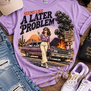 Puede incluir: Una camiseta de color morado claro con un estampado gráfico de una persona de pie ante un coche en llamas y humeante en un paisaje desértico. El texto dice SEEMS LIKE A LATER PROBLEM y SAME GIRL DIFFERENT DAY. Incluye un cactus.