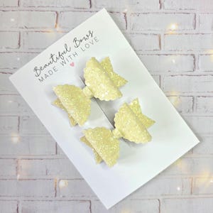 Pode incluir: Quatro laços de cabelo amarelos brilhantes em um cartão branco. Os laços são dispostos em um grupo, com um design de borda ondulada. O cartão tem o texto "Beautiful Bows MADE WITH LOVE" em escrita cursiva.