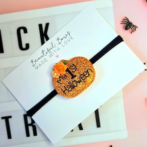Könnte beinhalten: Ein schwarzes elastisches Stirnband mit einem glitzernden orangefarbenen kürbisförmigen Ornament, das den schwarzen Text My 1st Halloween trägt. Das Ornament ist mit zwei kleinen orangefarbenen Stoffblumen verziert. Es ist auf einer weißen Karte befestigt.