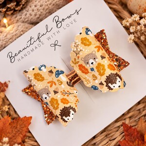 May include: A hair bow featuring a cream-coloured fabric with a pattern of bears, hedgehogs, mushrooms, and autumn leaves. The bow has scalloped edges and is accented with layers of orange glitter fabric. It is attached to a white display card.