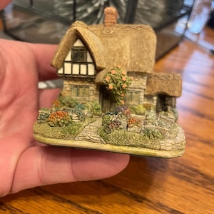 May include: A miniature cottage figurine with a thatched roof, brick chimney, and stone walls. The cottage has a small garden with colorful flowers and greenery. The figurine is held in a hand.