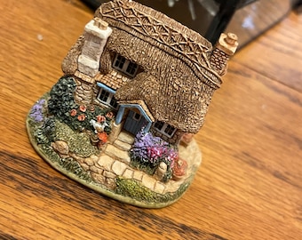 Może przedstawiać: Miniaturowa figurka domku z trzcinowym dachem, kamiennym kominem i kolorowymi kwiatami. Domek ma niebieskie drzwi i okna. Figurka stoi na drewnianej powierzchni.