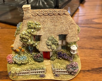 Może przedstawiać: Miniaturowa figurka domku z strzechą, czerwonymi drzwiami i małym białym płotem. Domek otoczony jest kolorowymi kwiatami i zielenią, tworząc uroczą, szczegółową scenę.