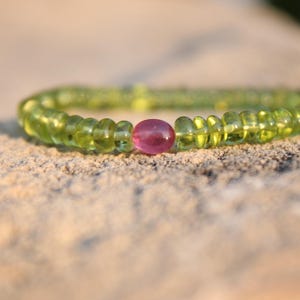 May include: A beaded bracelet featuring translucent, light green beads with a single, round, pink bead at the center. The bracelet is resting on a textured, light brown surface. The beads are evenly spaced.