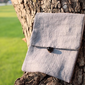 May include: A light gray linen clutch with a flap closure and a dark brown button. The clutch is set against a textured tree trunk, with a blurred green background. The bag is a simple, minimalist design.