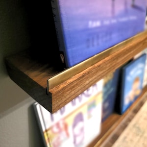 May include: A wooden floating bookshelf with a brass front edge. The shelf is filled with books, including one with a blue cover. The wood has a natural grain and a warm brown tone, creating a modern and minimalist design.