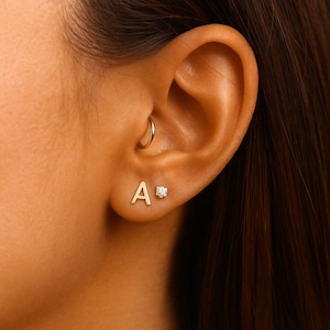 Könnte beinhalten: Nahaufnahme eines Ohrs mit zwei goldenen Ohrringen. Einer ist ein kleiner Buchstabe "A", der andere ein kleiner Stecker mit einem klaren Edelstein. Ein goldener Creolen-Ohrring ist ebenfalls sichtbar.