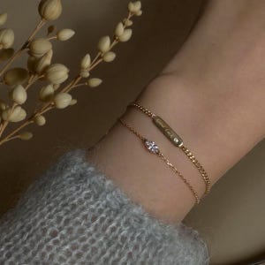 Könnte beinhalten: Zwei goldene Armbänder an einem Handgelenk. Eines ist eine zarte Kette mit einem kleinen, ovalen Edelstein. Das andere ist eine dickere Kette mit dem Schriftzug "Good Luck" auf einem rechteckigen Stab.