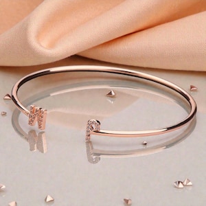May include: Rose gold open cuff bracelet with the letters "M" and "P" encrusted with small, clear crystals. The bracelet is displayed on a reflective surface with small, triangular decorative accents.