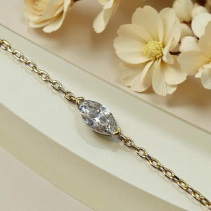 Puede incluir: Una pulsera de cadena dorada con un diamante talla marquesa. El diamante est&aacute; engastado en un bisel de oro. La cadena tiene un dise&ntilde;o de eslabones. La pulsera se exhibe sobre una superficie blanca. El diamante es el punto focal.