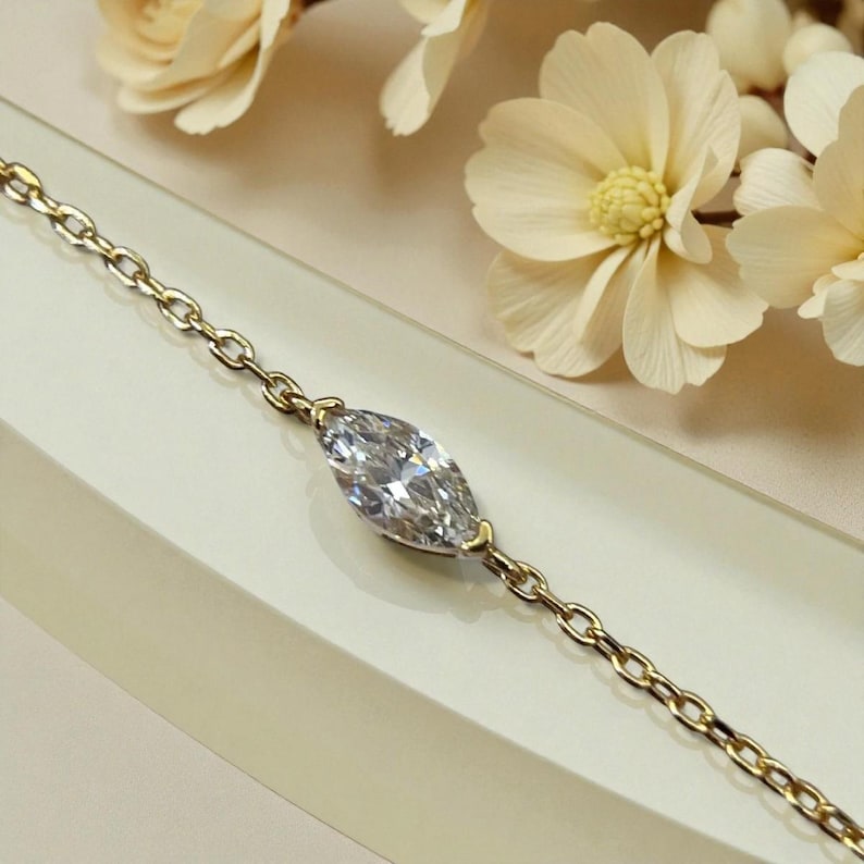 Puede incluir: Una pulsera de cadena dorada con un diamante talla marquesa. El diamante est&aacute; engastado en un bisel de oro. La cadena tiene un dise&ntilde;o de eslabones. La pulsera se exhibe sobre una superficie blanca. El diamante es el punto focal.