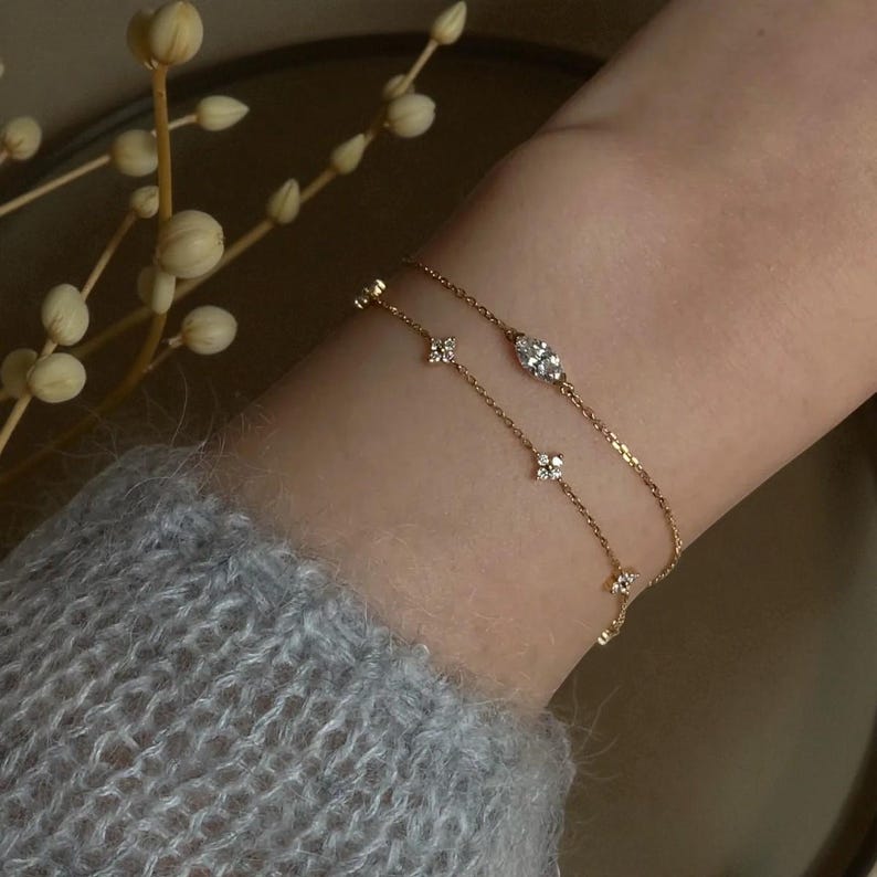 Puede incluir: Dos delicadas pulseras de oro con brillantes detalles de diamantes se muestran en una mu&ntilde;eca. Una pulsera presenta un diamante ovalado central, mientras que la otra tiene peque&ntilde;os racimos de diamantes en forma de flor. Las pulseras est&aacute;n superpuestas, creando un aspecto sutil y elegante.