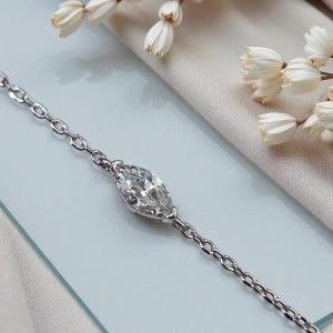 Puede incluir: Una delicada pulsera de plata con un diamante talla marquesa como pieza central. El diamante est&aacute; engastado en una montura de garras y est&aacute; conectado a una fina cadena. La pulsera se muestra sobre una superficie azul claro, con flores blancas en el fondo.