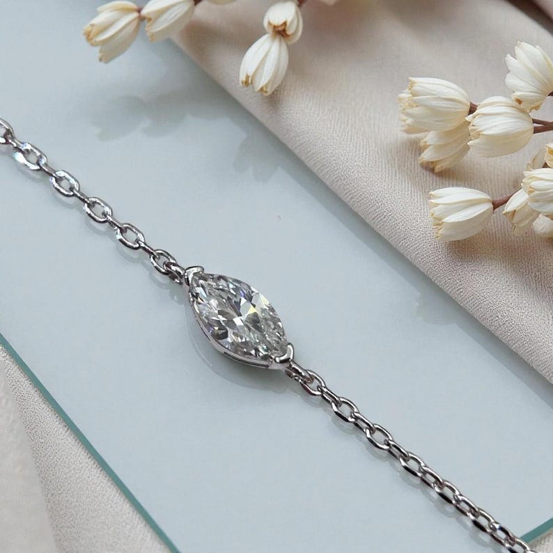 Puede incluir: Una delicada pulsera de plata con un diamante talla marquesa como pieza central. El diamante est&aacute; engastado en una montura de garras y est&aacute; conectado a una fina cadena. La pulsera se muestra sobre una superficie azul claro, con flores blancas en el fondo.