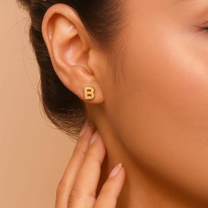 Może przedstawiać: Zbliżenie ucha z kolczykiem w kształcie litery "B" w kolorze złota. Kolczyk jest mały i kwadratowy. Odcień skóry jest jasny, a tło ma miękki, neutralny kolor.