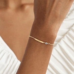 Könnte beinhalten: Ein goldfarbenes Armband mit einer filigranen Kette und einem rechteckigen Steg mit dem eingravierten Namen "Bobbi". Ein kleiner, runder, hellblauer Edelstein ist in der Nähe des Verschlusses gefasst. Das Armband wird am Handgelenk getragen.