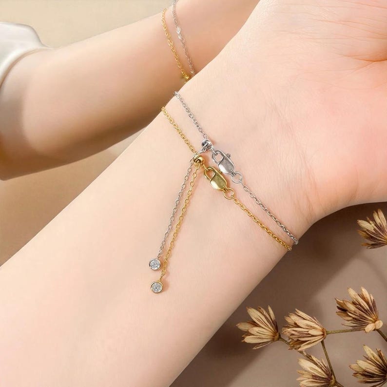 Puede incluir: Dos pulseras delicadas, una dorada y otra plateada, adornan una mu&ntilde;eca. Cada pulsera presenta una fina cadena con un peque&ntilde;o dije colgante. Una pulsera tiene un cierre dorado y la otra un cierre plateado. Las pulseras est&aacute;n adornadas con peque&ntilde;as gemas brillantes.