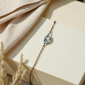 Puede incluir: Una delicada pulsera de cadena dorada con una gema transparente de talla marquesa. La pulsera se exhibe sobre una superficie color crema con una tela de tonos neutros en el fondo. La joyer&iacute;a es sencilla y elegante.