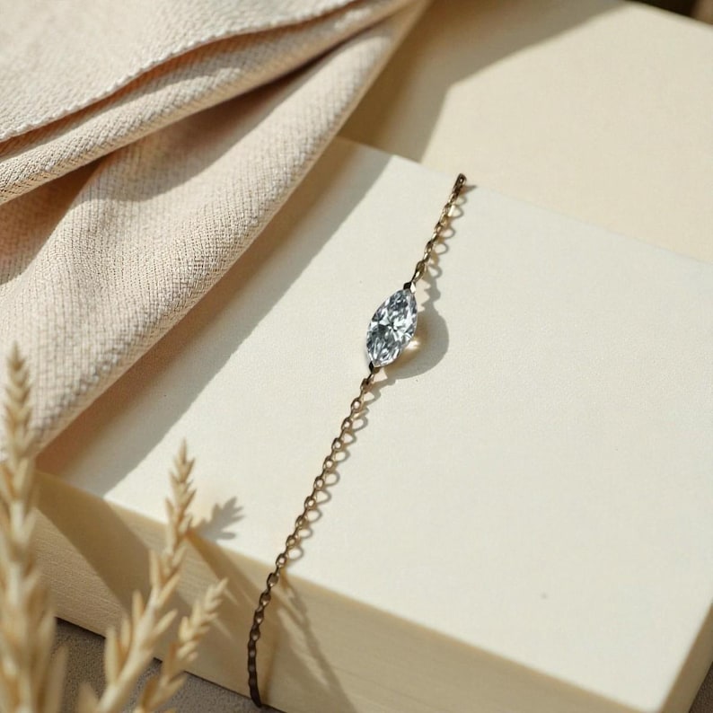 Puede incluir: Una delicada pulsera de cadena dorada con una gema transparente de talla marquesa. La pulsera se exhibe sobre una superficie color crema con una tela de tonos neutros en el fondo. La joyer&iacute;a es sencilla y elegante.