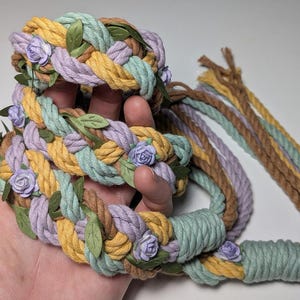 May include: Braided rope headbands in pastel colors of yellow, lavender, and mint green, accented with small purple flowers and green leaves. The headbands are handmade and feature a rustic, bohemian style.