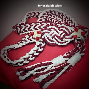 May include: A handcrafted, decorative rope piece in burgundy and white, featuring a braided design and adorned with small red and cream-colored roses. The text "Personalizable colors!" is visible.