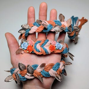 May include: A braided cord accessory featuring intertwined teal, peach, and light pink fabric strands. The braid is decorated with small, light blue and orange artificial roses and metallic silver-toned leaf charms attached along the length.