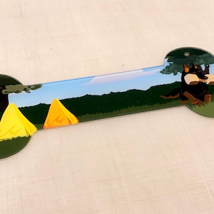 May include: Bone-shaped decorative sign with a camping theme. The sign features a cartoon dog, two yellow tents, a tree, and a blue sky background. The dog is black and brown. The tents are yellow and orange.