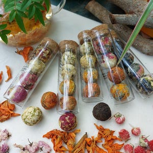 May include: Assortment of colorful tea balls in clear glass tubes with cork stoppers. The tea balls are various colors, including pink, yellow, and orange. Dried flowers and a glass teapot are also visible.