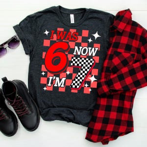 Puede incluir: Camiseta gris oscuro con el texto "I WAS 6 NOW I'M 7" en rojo y blanco, con un dise&ntilde;o a cuadros. Una camisa a cuadros roja y negra est&aacute; doblada junto a la camiseta. Botas negras y gafas de sol tambi&eacute;n est&aacute;n en la imagen.