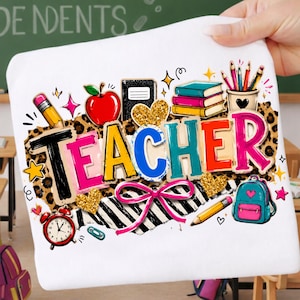 Könnte beinhalten: Ein weißes T-Shirt mit einer bunten Grafik und dem Wort TEACHER in fetten Buchstaben. Das Design zeigt einen roten Apfel, einen Bücherstapel, einen Stiftehalter, einen Rucksack, einen Wecker, goldene Glitzerherzen und eine rosa Schleife auf Leopardenmuster.